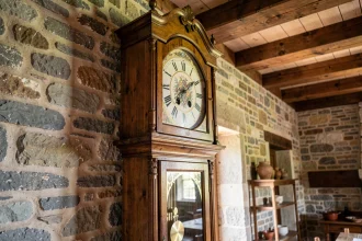 Horloge comtoise ancienne avec cadran fleuri et balancier doré contre un mur en pierre d'une maison de campagne française