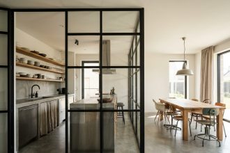Cuisine ouverte avec verrière industrielle en acier noir séparant le coin repas, plan de travail en béton et étagères en bois